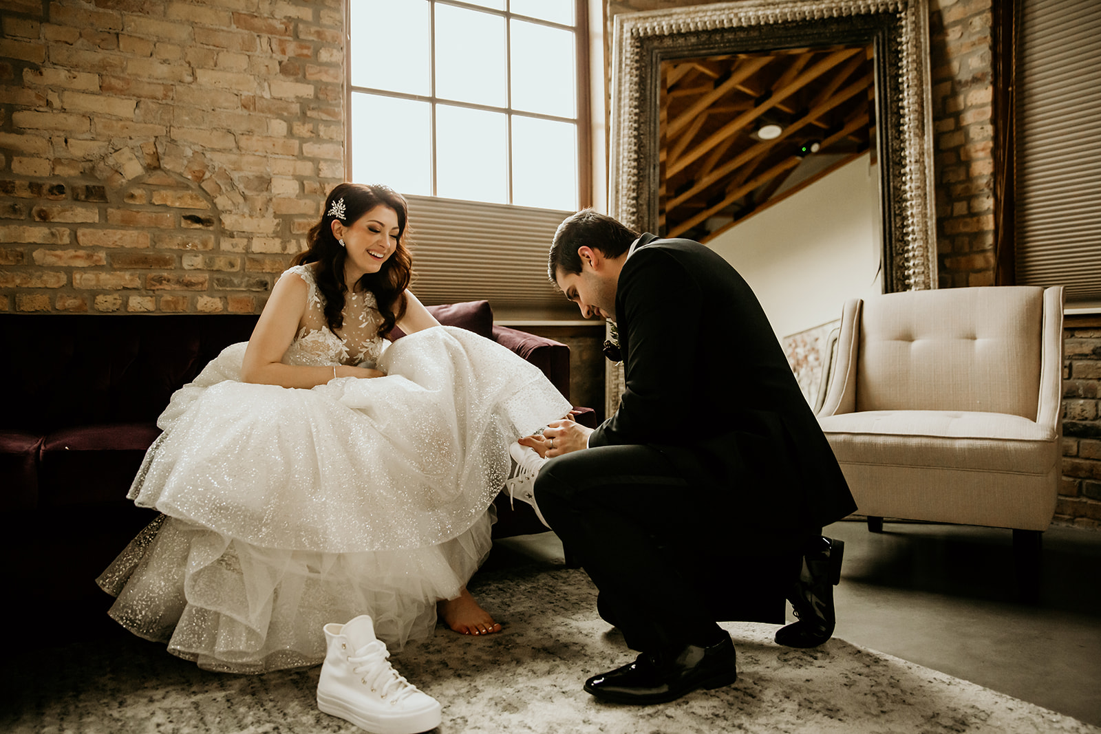 Groom helping his bride switch from heels to sneakers in an elegant bridal room.