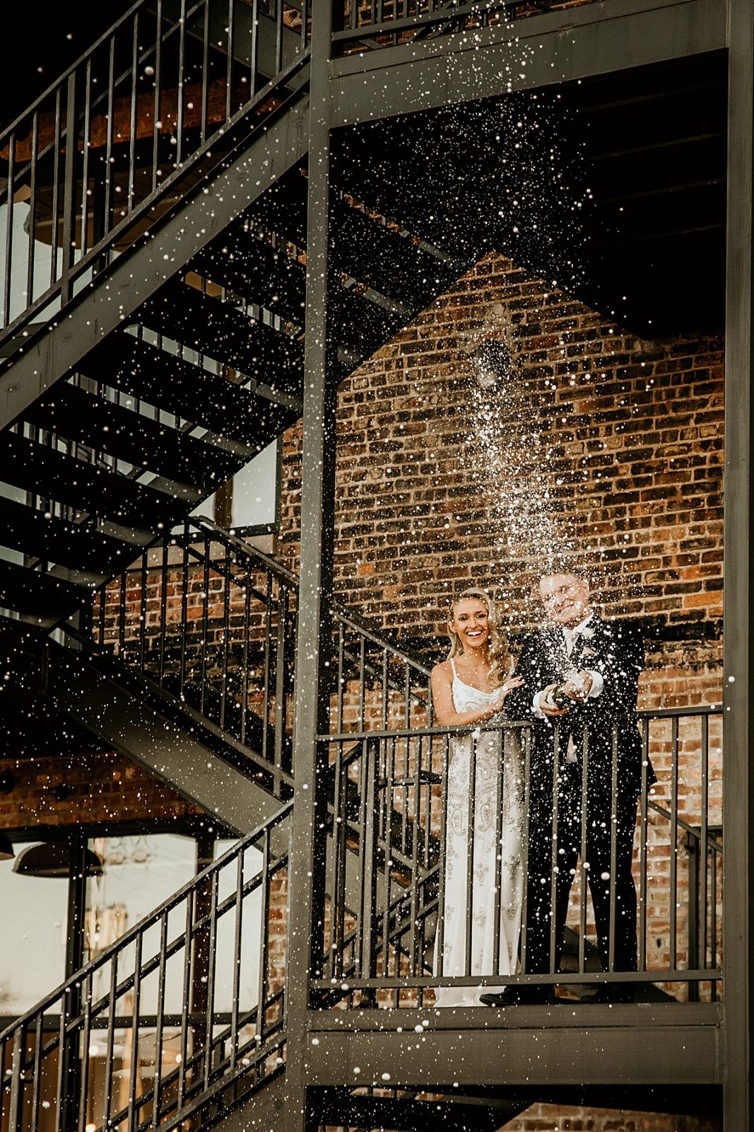 A bride and groom popping a bottle of champagne off an outdoor staircase