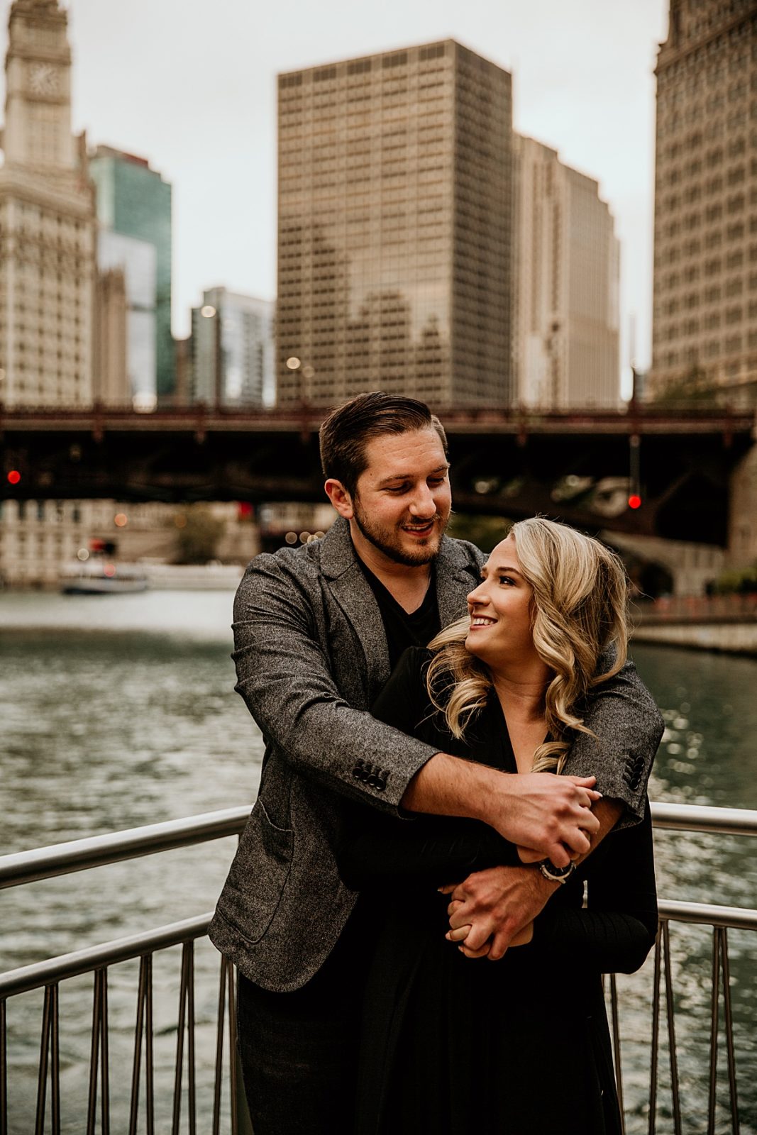 a man hugging his fiancé from behind while they look at each other on the chicago river walk with the city behind them
