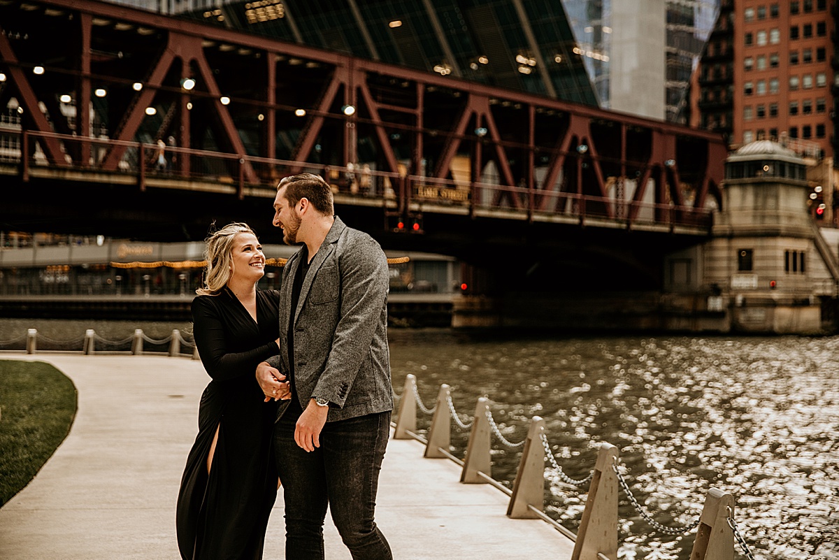 a young couple holding hands, walking on the chicago river walk with a bridge behind them