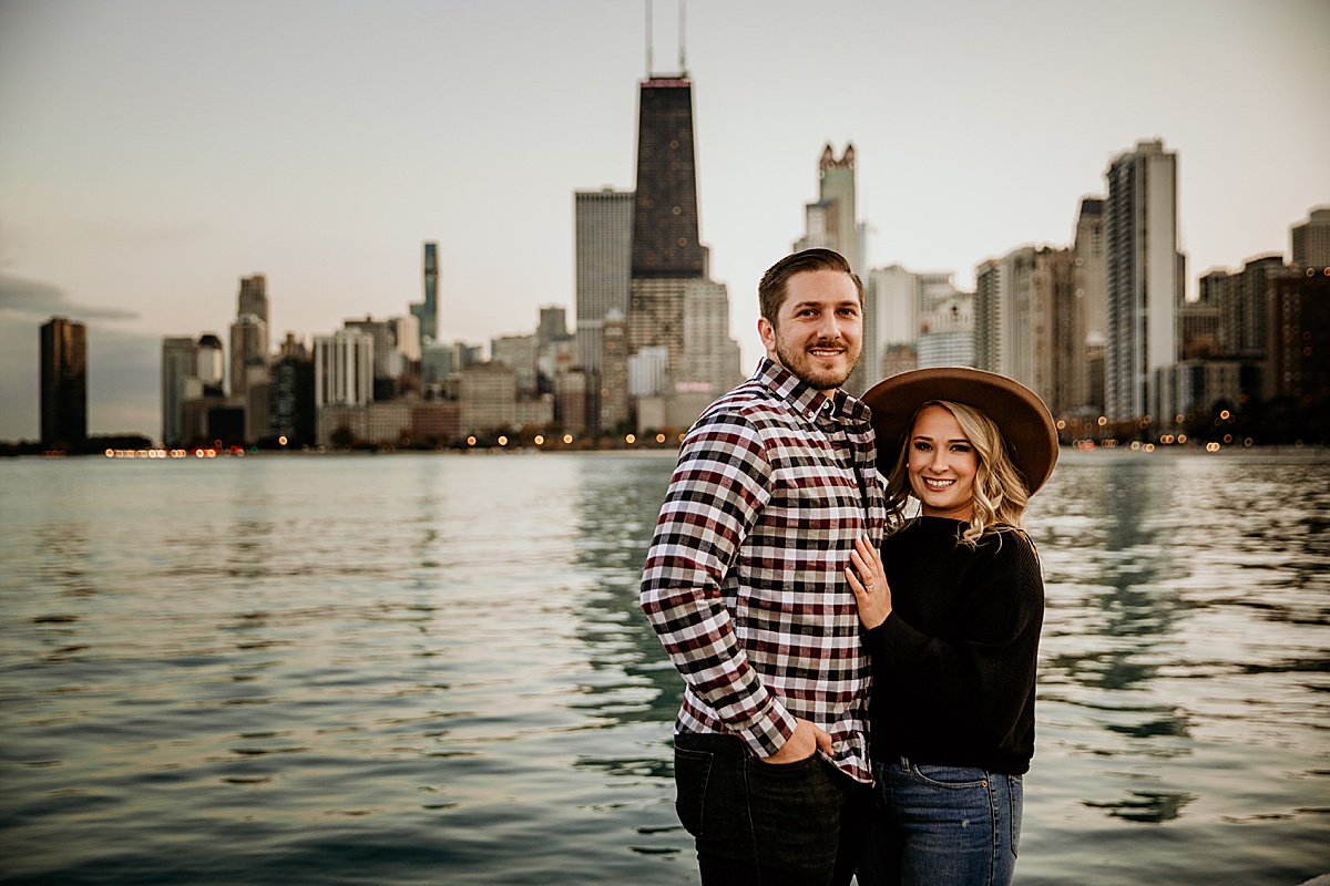 A young engaged couple looking at the camera with the chicago skyline behind them at north ave beach during the sunset. 