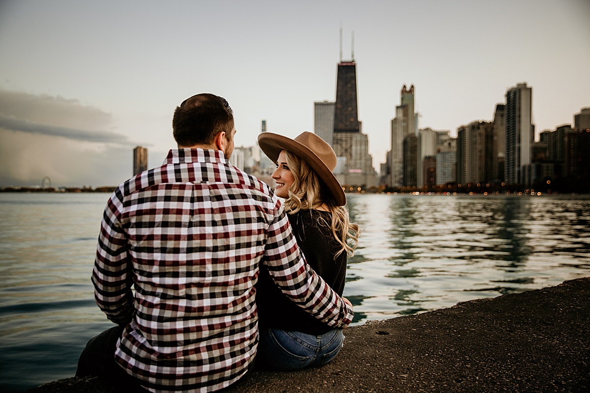 A young engaged couple looking at each other, sitting on the edge of lake Michigan with the Chicago skyline behind them at north ave beach during the sunset. 