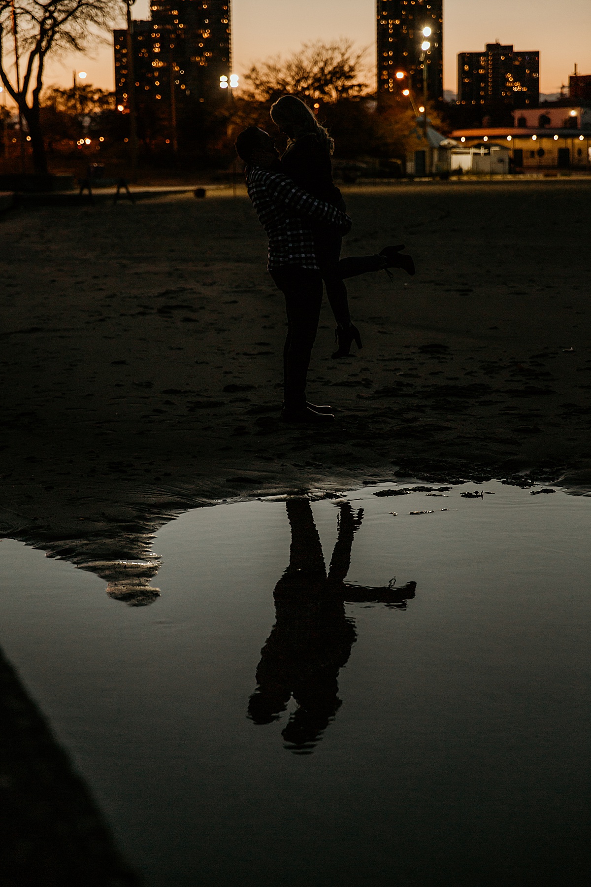 A reflection of man lifting his fiancé up in a large puddle of water from Lake Michigan at sunset with the city of chicago behind them.