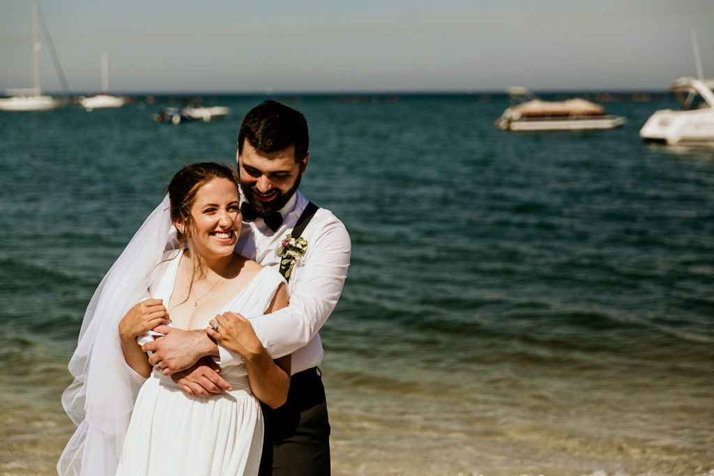 Groom hugging bride from behind on Lake Michigan beach.